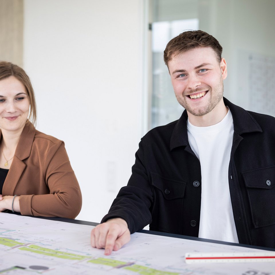 Zwei junge Mitarbeiter mit Bauplänen am Tisch – Szenenfoto für Karriereseite.